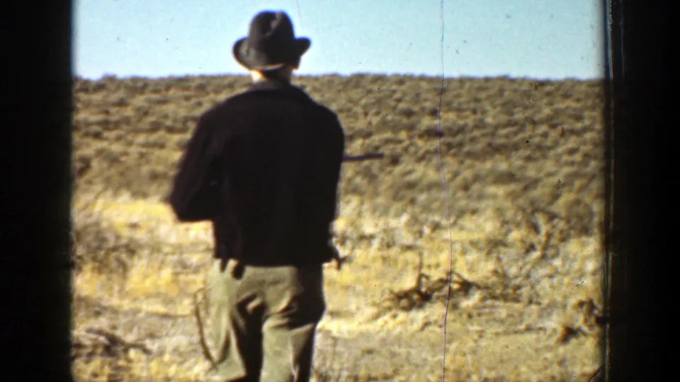 1936 California 8mm frame of a man in a dark jacket and hat walking away through dry brush with a rifle at his side.