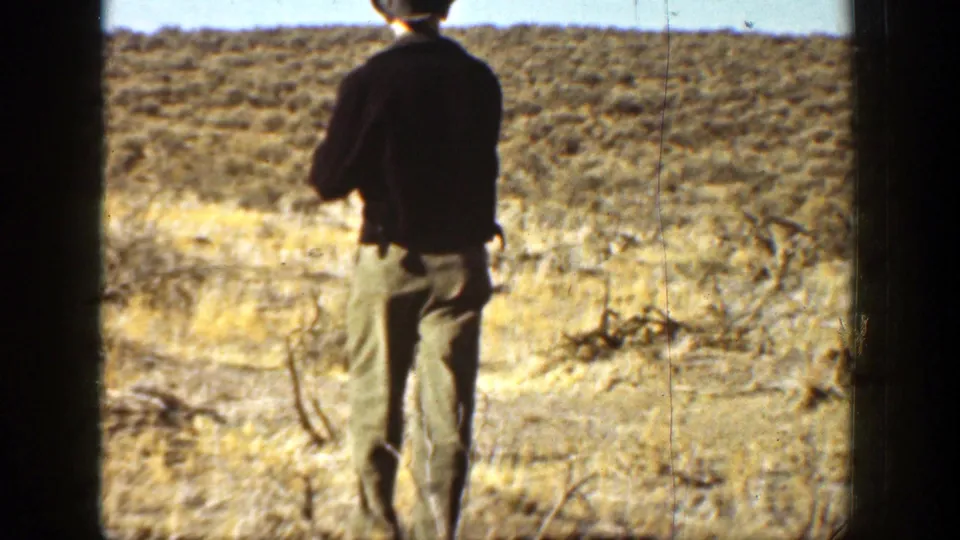 1936 California home-movie frame showing a man from behind in a dark jacket walking through scrubland with a rifle.