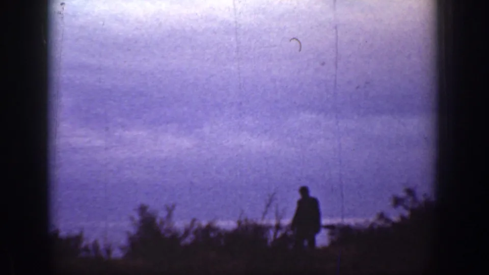 1936 California 8mm frame of a dark silhouetted figure beside hillside brush, with a purple dusk sky or water surface behind.