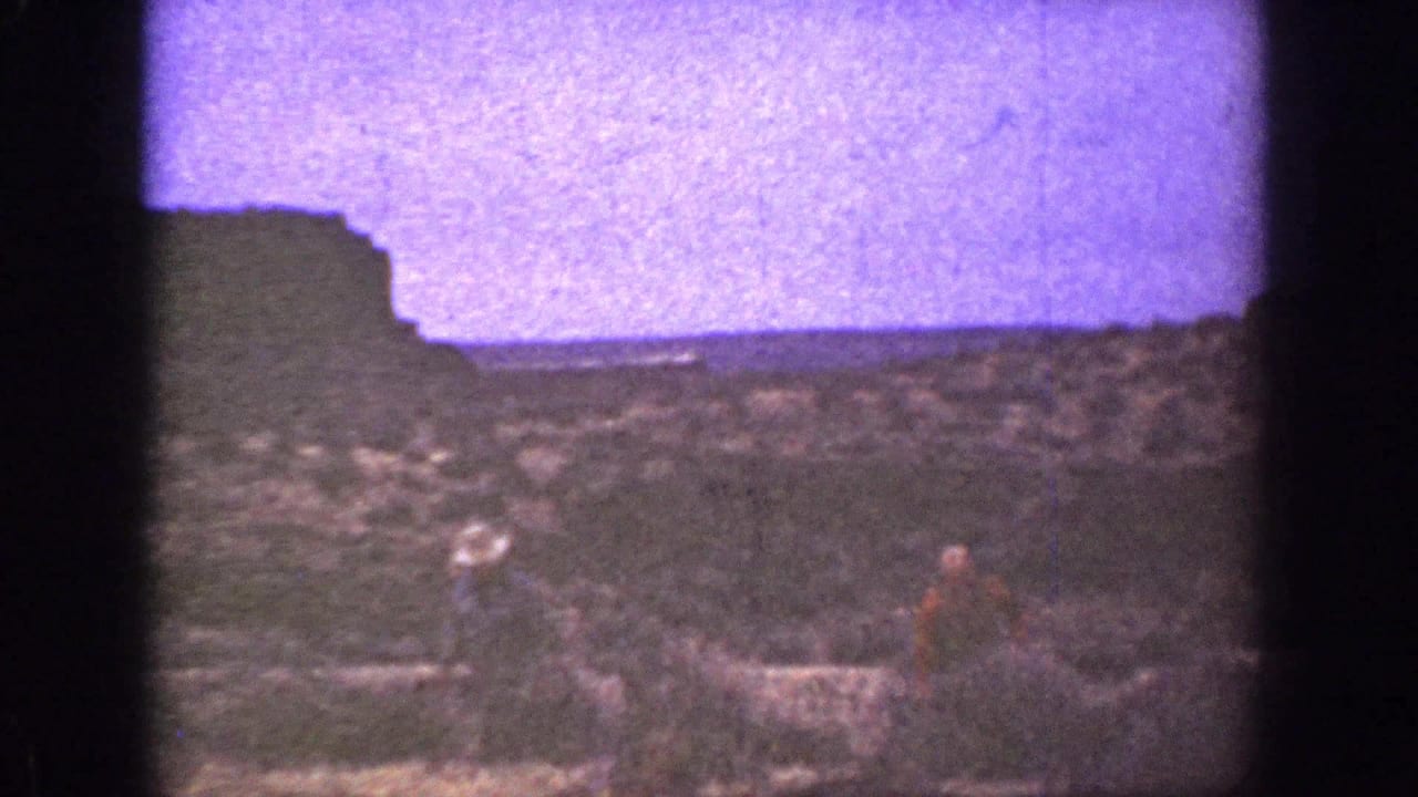 1936 California 8mm frame showing two men in hats crossing rough desert ground near a dark bluff and hazy horizon.