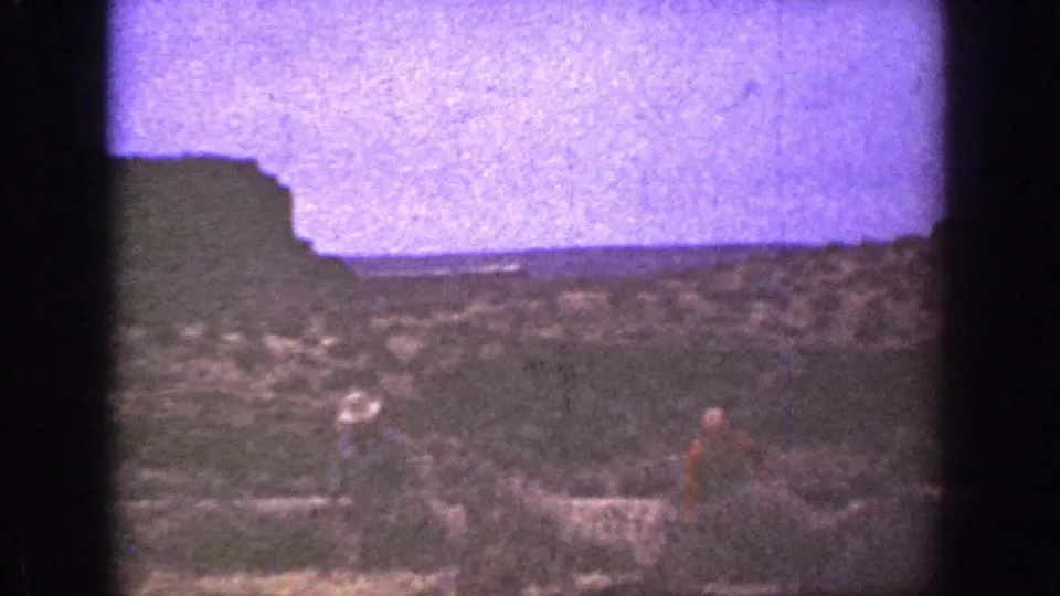 1936 California 8mm frame showing two men in hats crossing rough desert ground near a dark bluff and hazy horizon.