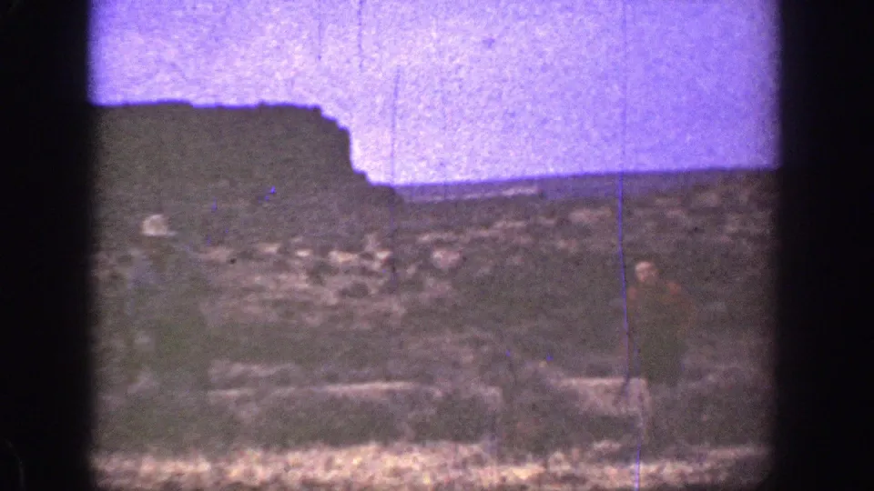 Grainy 1936 California home-movie frame showing two men on scrubby desert ground below a dark rock formation.