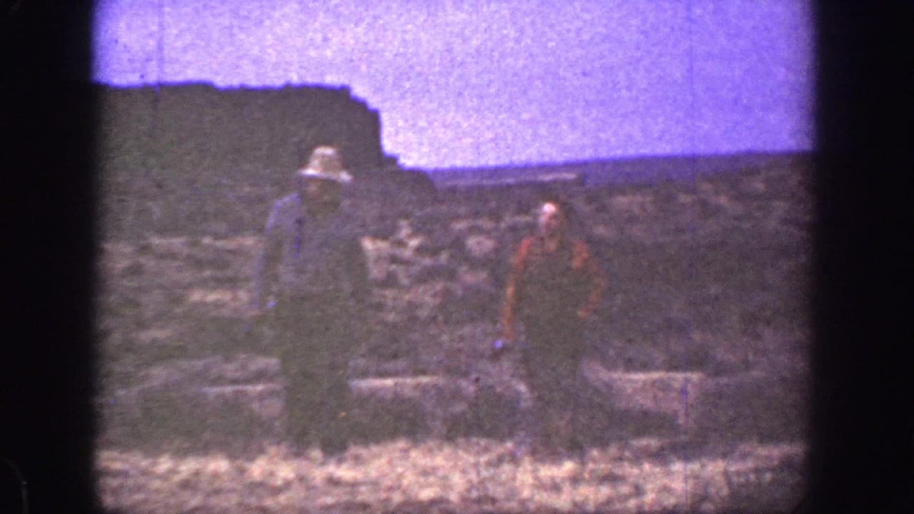 Two men walk across dry rocky ground beneath a purple-tinged sky.