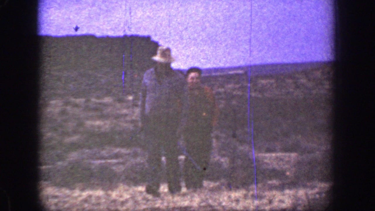 Two men continue walking toward the camera with a rocky slope behind them.
