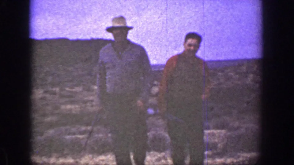 Two adult men in vintage 8mm footage walk side by side in a dry California landscape, one carrying an indistinct object.