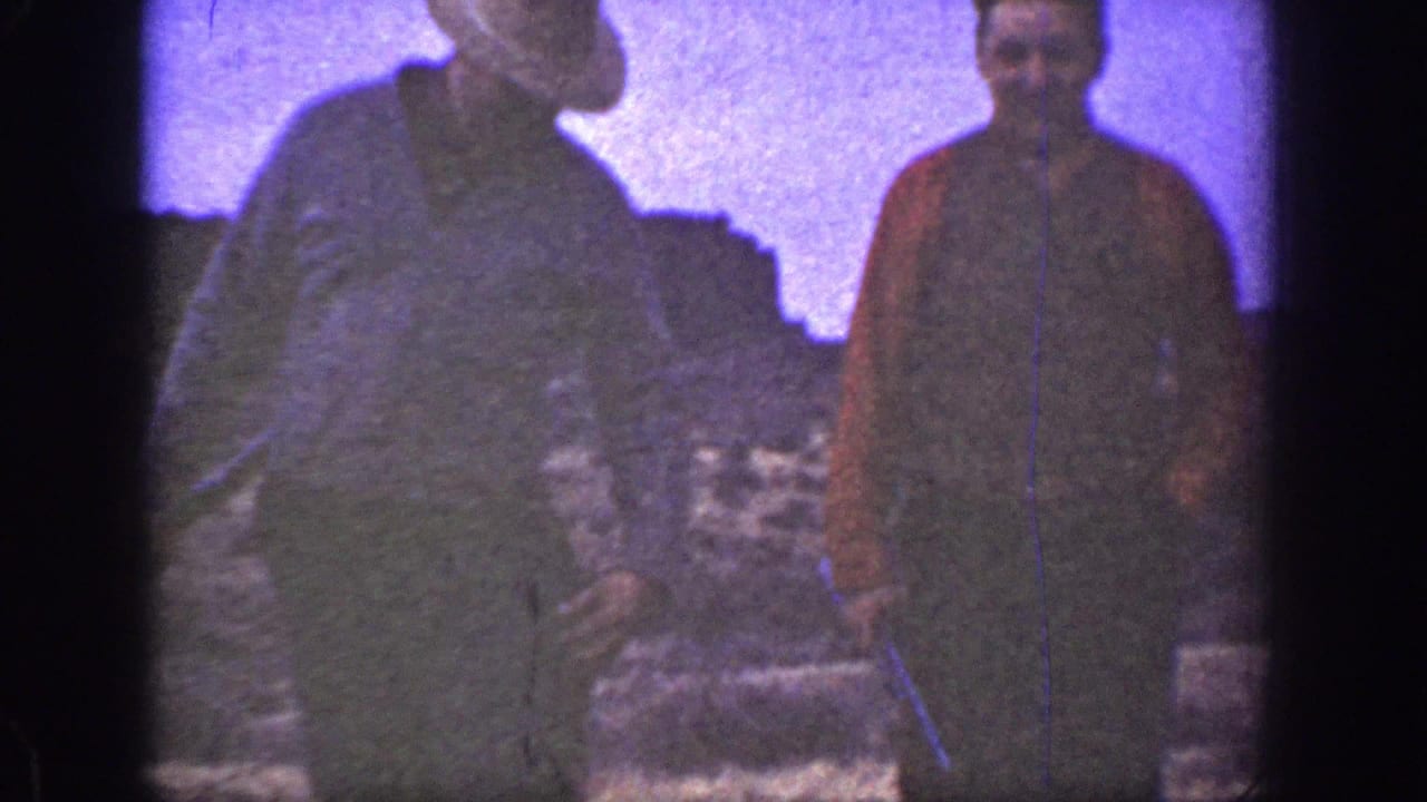 Two adult men in 1936 California home-movie footage stand on arid terrain, one wearing a light hat and the other a red shirt.