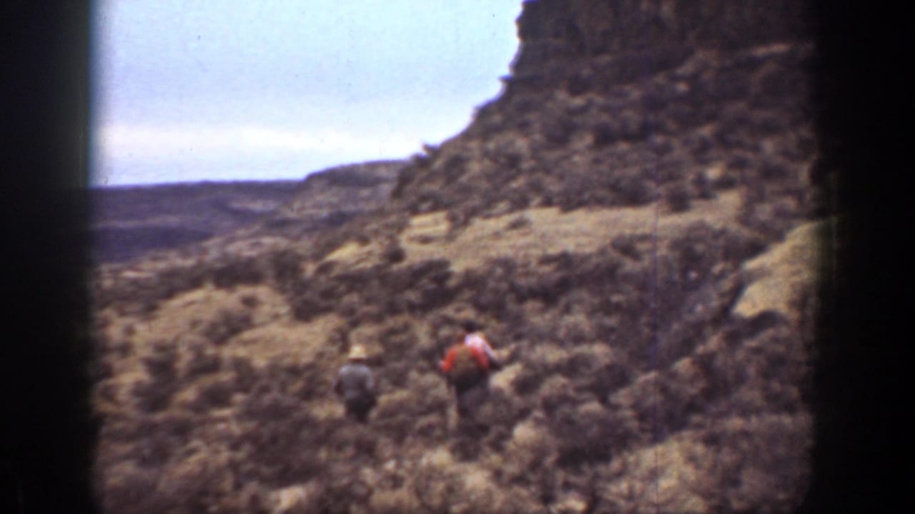 Three men hike across a rocky, brush-covered slope below a steep hillside.