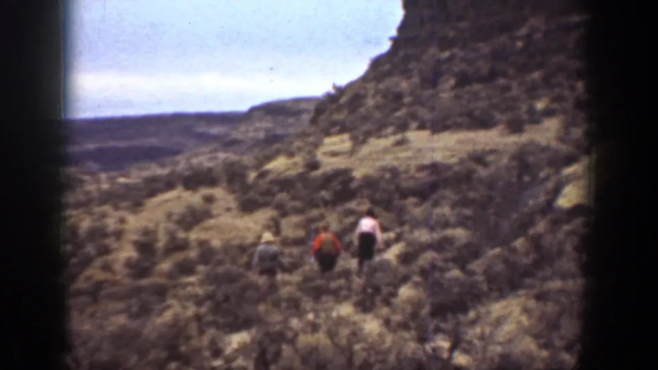 Three men hike away across a rugged California slope, with dry vegetation, rocky ground, and a dark 8mm frame edge visible.