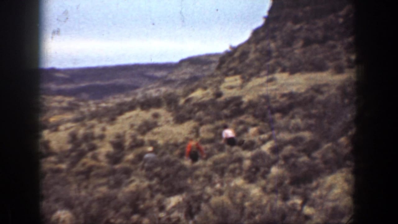 1936 California 8mm home-movie frame showing distant hikers crossing brushy canyon terrain beneath a steep rocky slope.