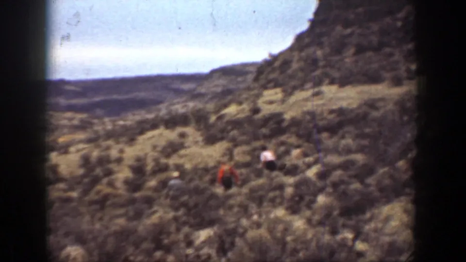 1936 California 8mm home-movie frame showing distant hikers crossing brushy canyon terrain beneath a steep rocky slope.