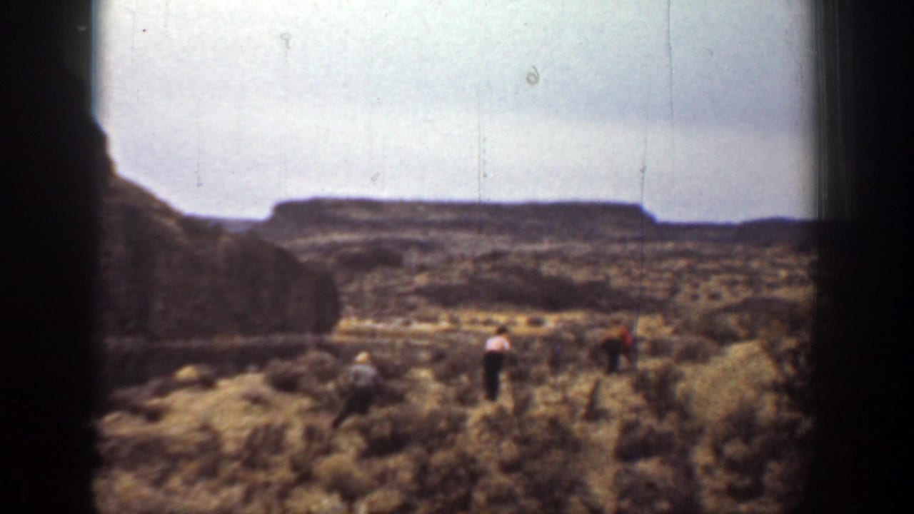 Hikers spread across brushy terrain with flat ridges in the distance.