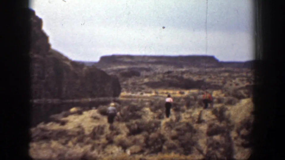 1930s home-movie view of hikers navigating brush-covered canyon terrain in California, framed by rock walls and distant high ground.