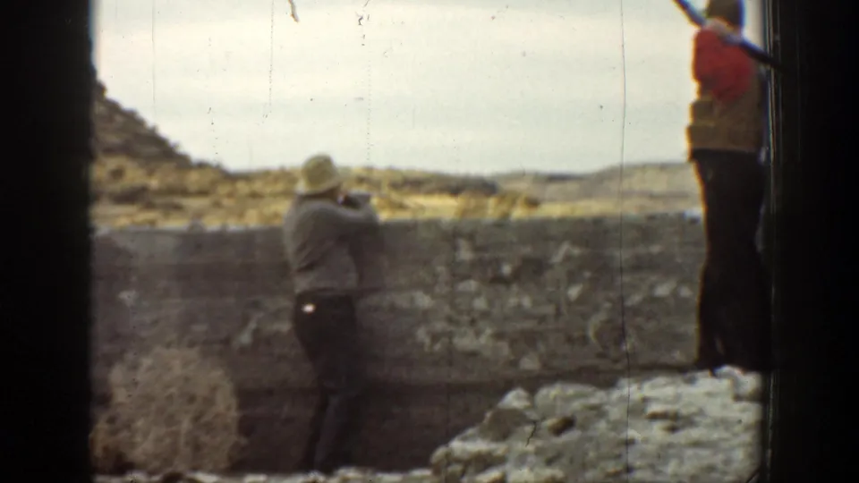 1936 California 8mm frame showing two adult men near a stone wall, with one man in a hat holding a rifle and another partly visible on the wall.