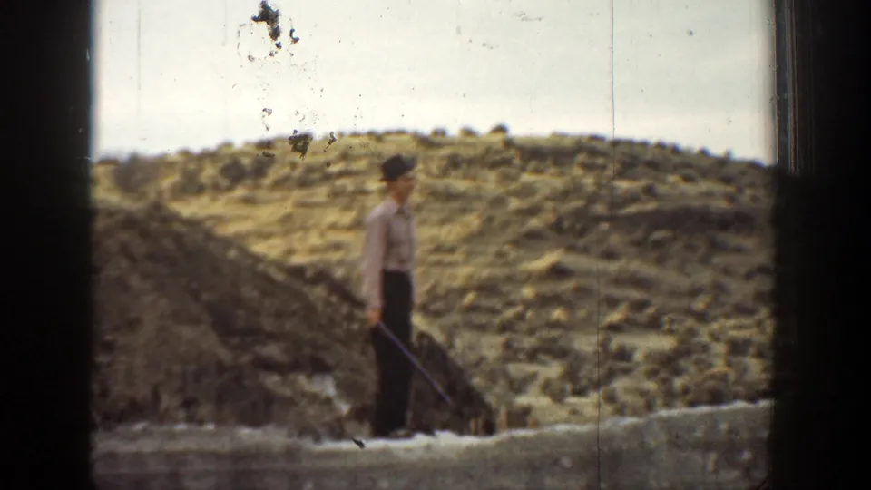 1936 California 8mm frame showing an adult man holding a rifle while standing near rocks and dry low hills.