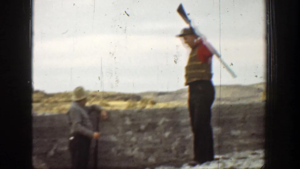 Two men in period outdoor clothing handle long guns in a dry California landscape, one standing at right with a gun over his shoulder.