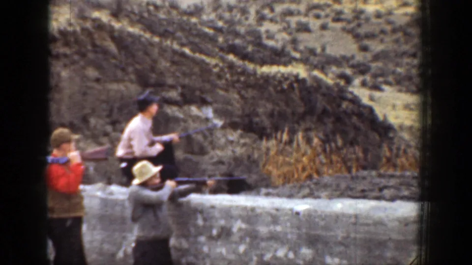 Men with long guns stand and aim behind a low stone wall in 1930s California home-movie footage, with scrubby hills behind them.