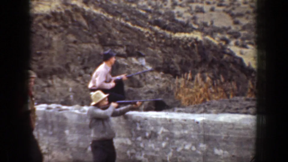 Two men in hats handle long guns from behind a low wall in a dry California landscape, with rugged hills in the background.