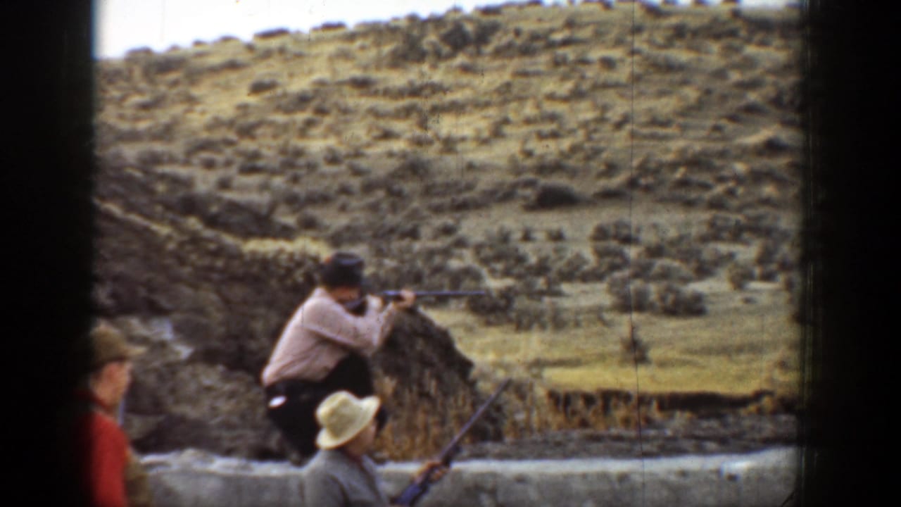 1936 California home-movie frame showing men with rifles near a low stone wall in a scrub-covered canyon landscape.