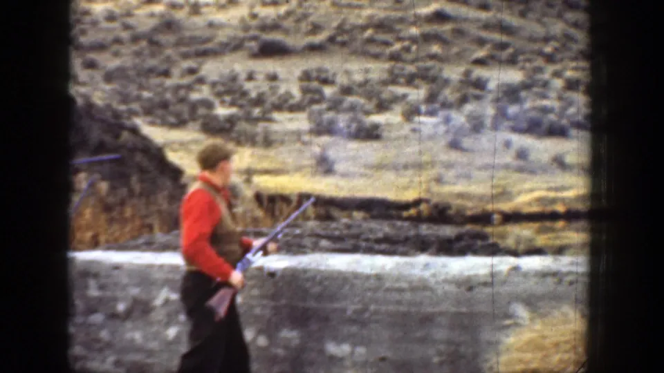 1936 California home-movie frame showing a man in a red sweater and vest holding a rifle near a canyon stone wall.