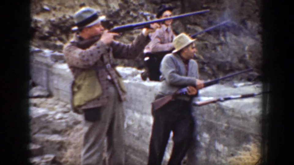 1936 California home-movie frame showing men in hats aiming and holding rifles behind a stone wall in a canyon.