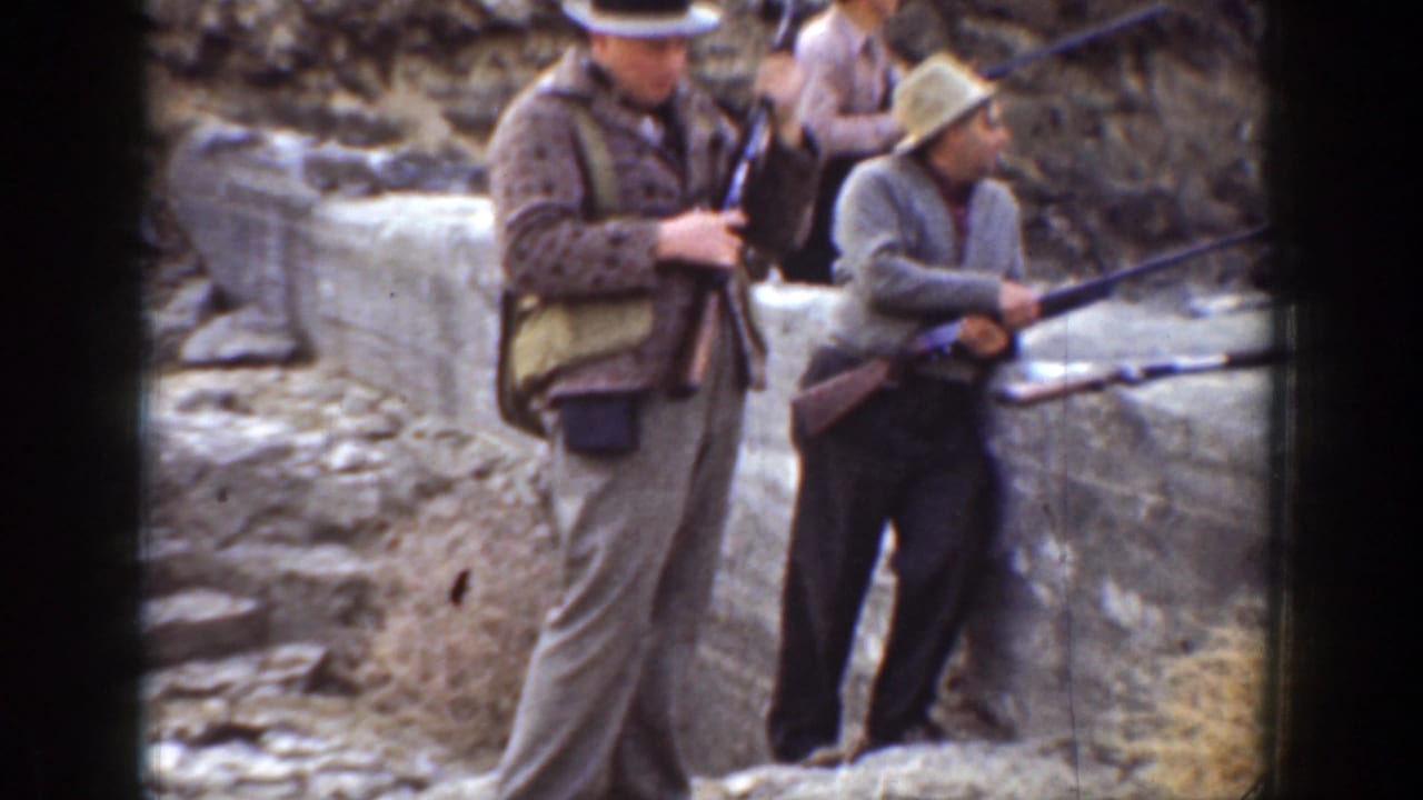 1936 California home-movie frame of three men in hats and jackets standing by grey rocks with rifles.