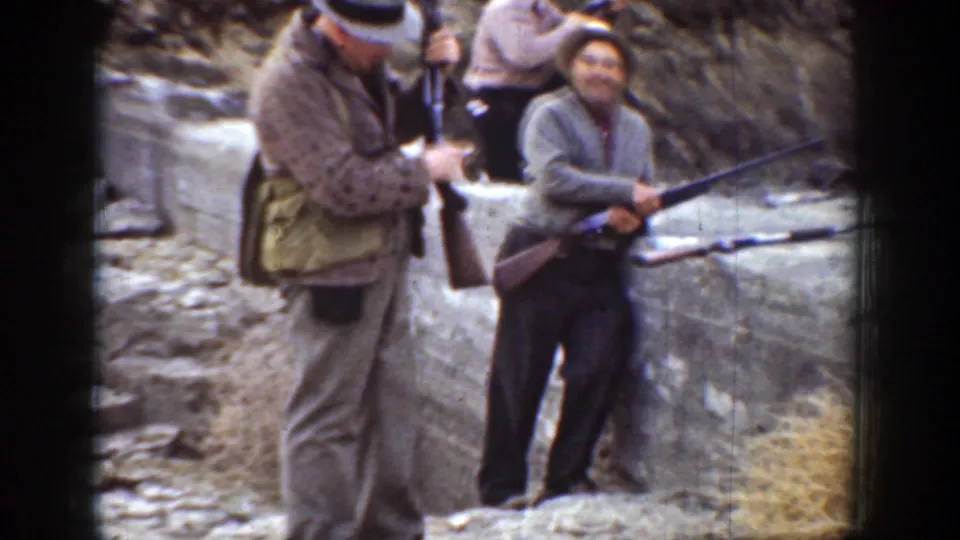 1936 California home-movie frame with men in period outdoor clothing holding rifles beside grey rocks.