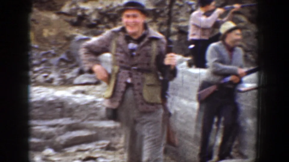 1936 California frame of a smiling man in outdoor clothing beside another man holding a rifle near rocks.