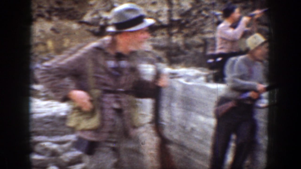 Man in a brimmed hat holds a shotgun near a rocky embankment while other men stand behind him.