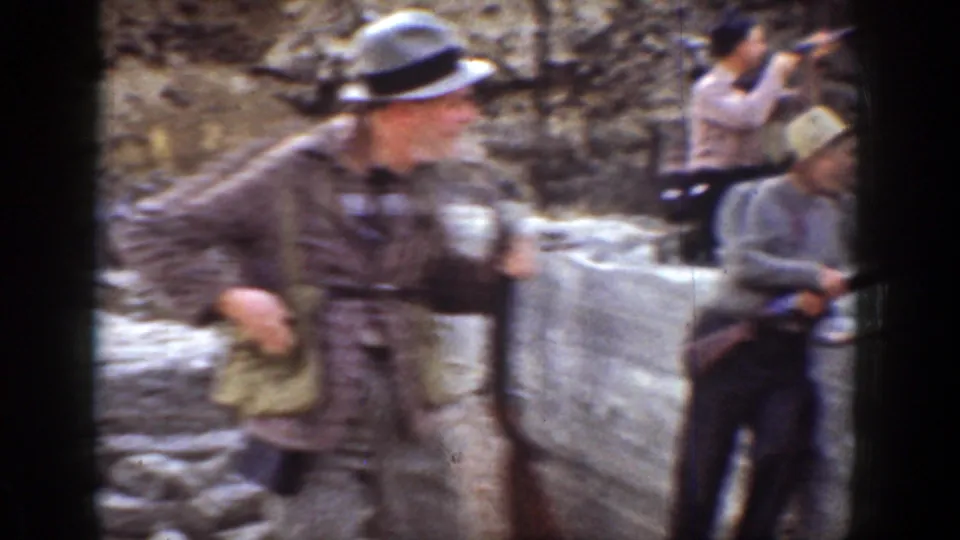 1936 California 8mm frame of men with shotguns near a rocky arid embankment, including a foreground man in a hat and patterned jacket.
