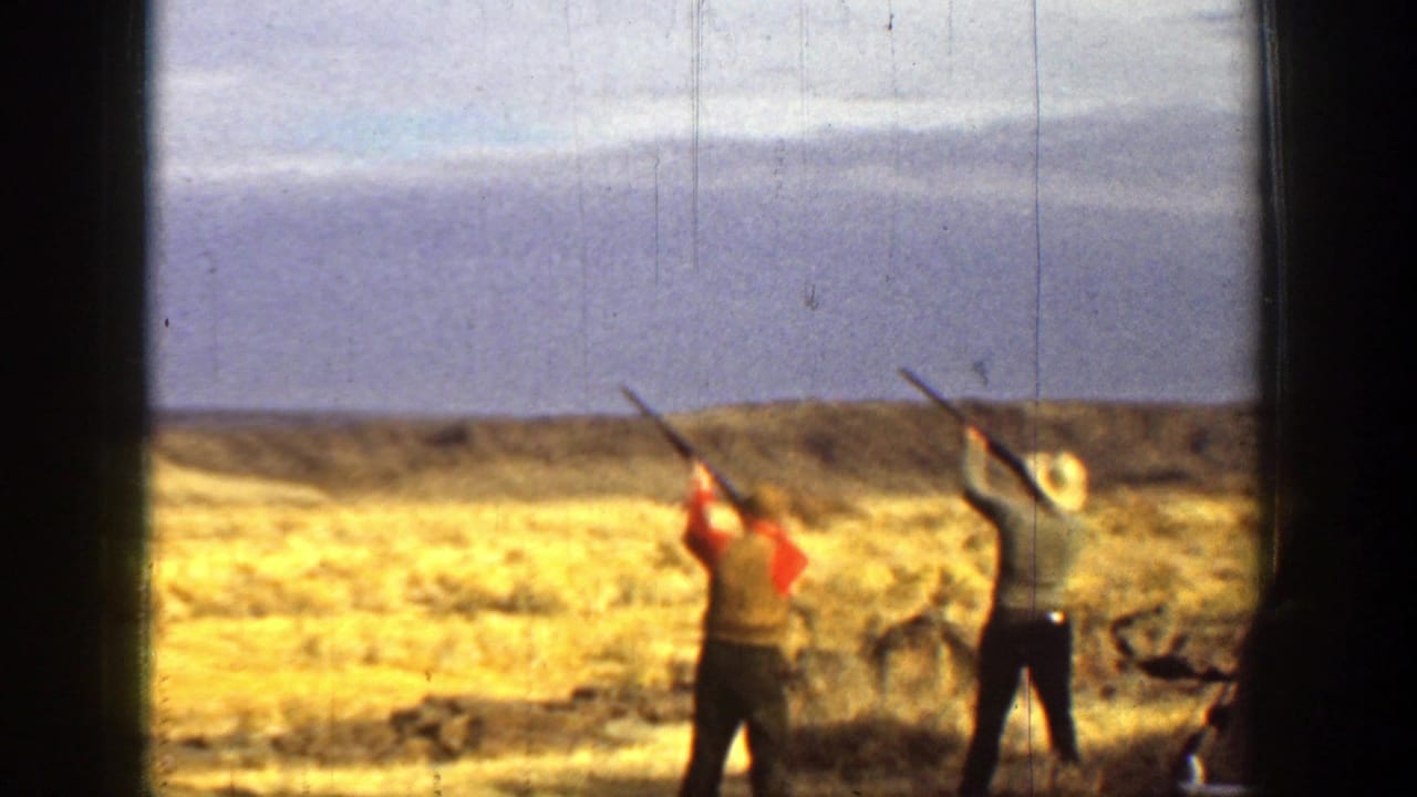 Two men raise shotguns toward the sky in an open dry field with low hills behind them.