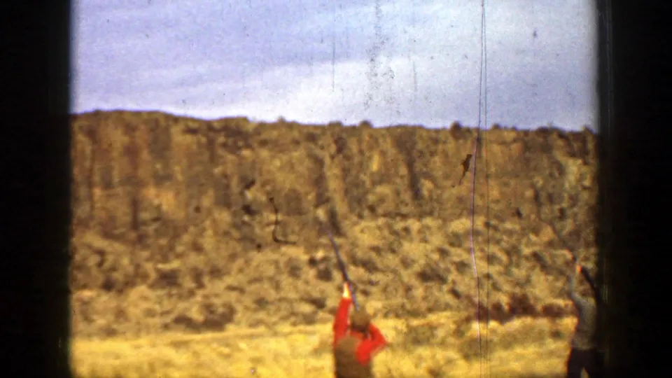 1936 California 8mm frame of men with raised shotguns below a rocky hillside during clay pigeon shooting.