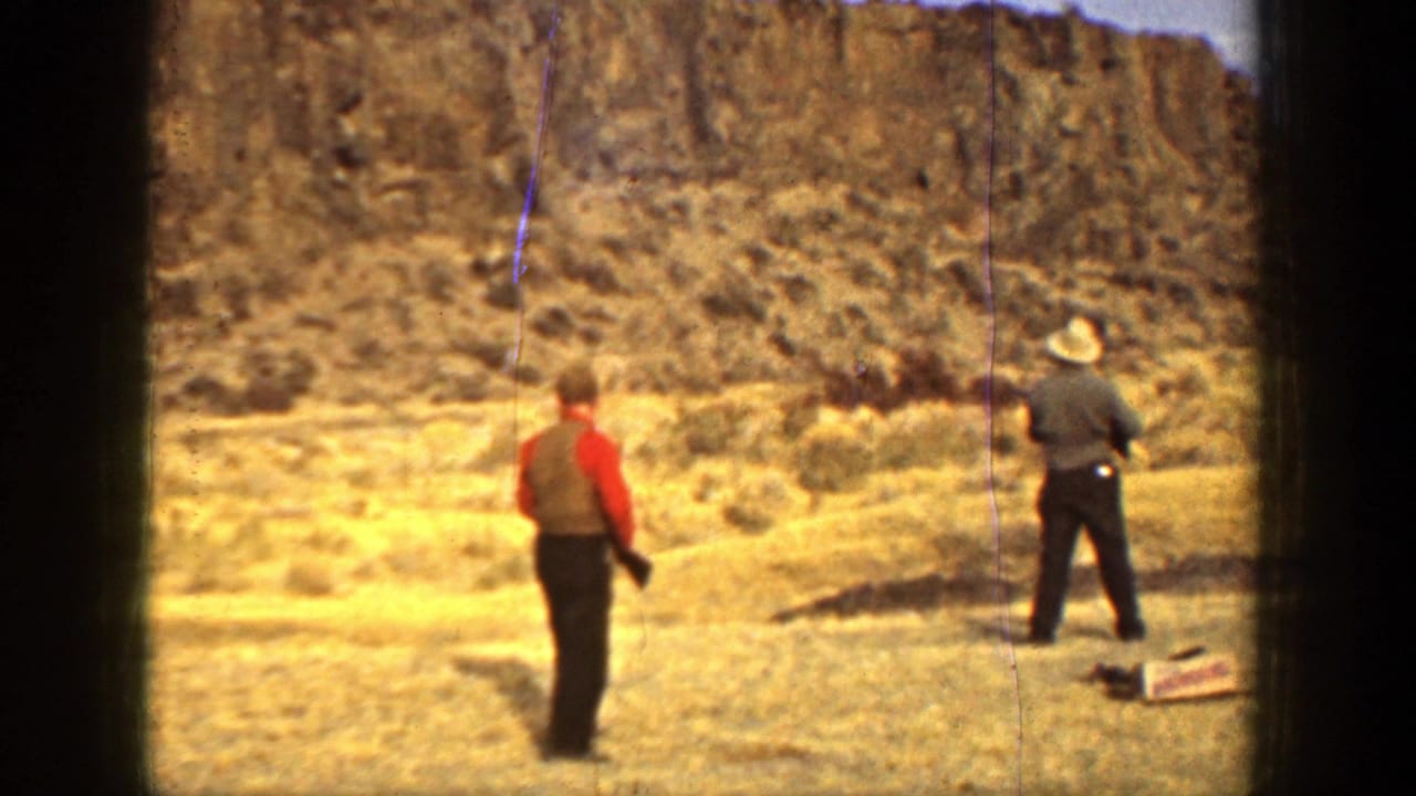 Two adult men stand in a dry field below a rocky cliff, one in red and one in a light hat.