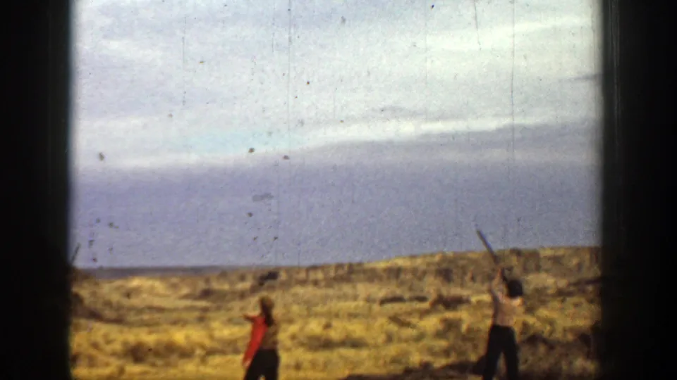 Two men in a 1936 California dry landscape raise their arms toward the sky, framed by open sky, brush, and soft 8mm film texture.