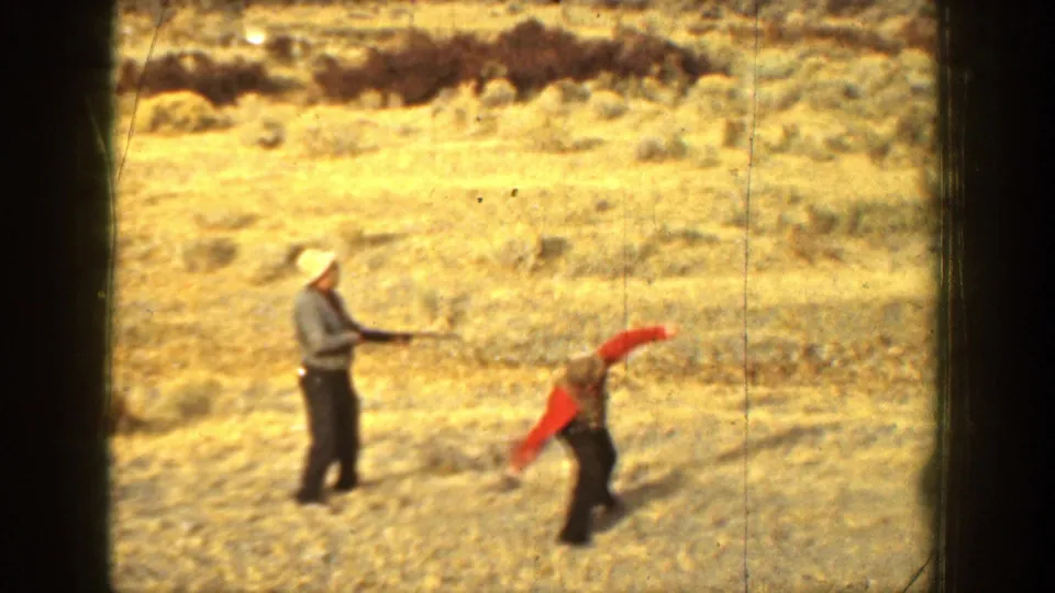 Two men are visible in a dry California field, with one holding a shotgun and the other bending or stepping as the skeet practice continues.