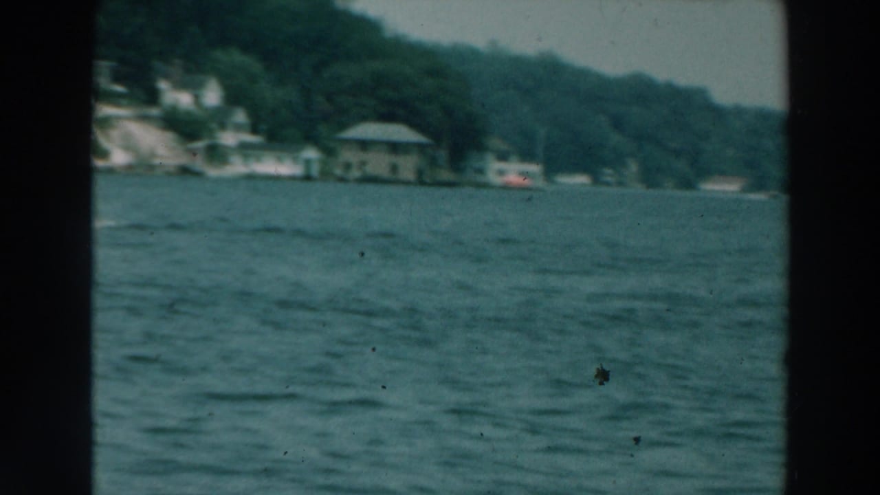 Lake water seen from a boat with wooded shoreline and small lakeside buildings in the distance.