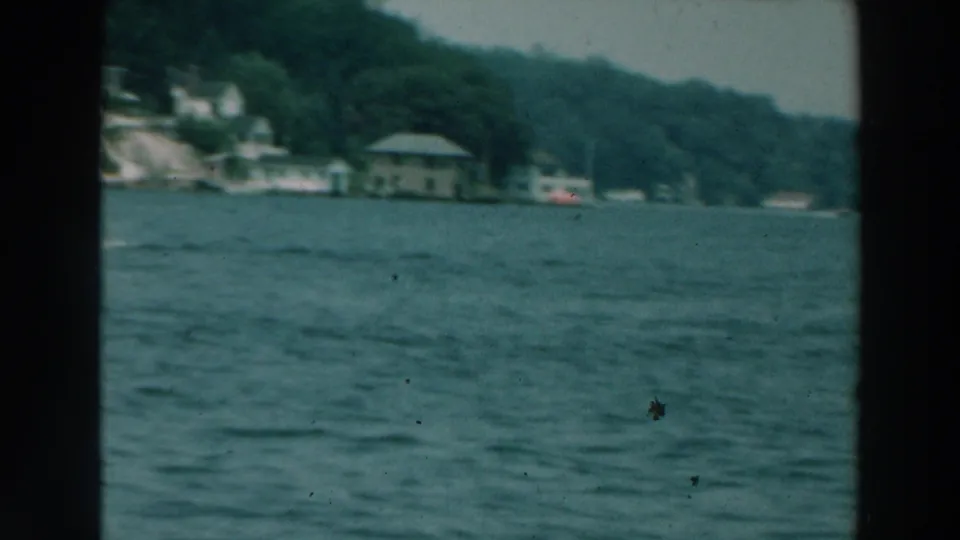 1964 Lake Winnebago home-movie frame showing rippling water, distant wooded shore and scattered buildings from a moving boat.