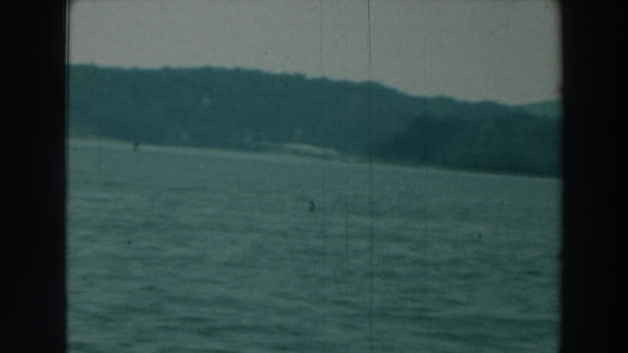 1964 8mm view of Lake Winnebago with rippled water, dark gate borders, hazy sky, and a tree-lined shore across the horizon.