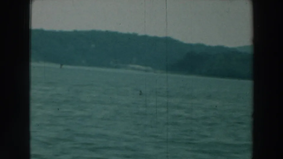 1964 8mm view of Lake Winnebago with rippled water, dark gate borders, hazy sky, and a tree-lined shore across the horizon.