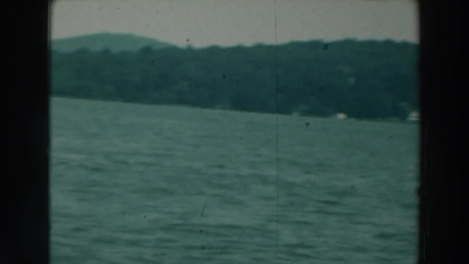 1964 Lake Winnebago footage showing open rippled water, a low tree-covered shore, muted sky, and visible 8mm film edges.