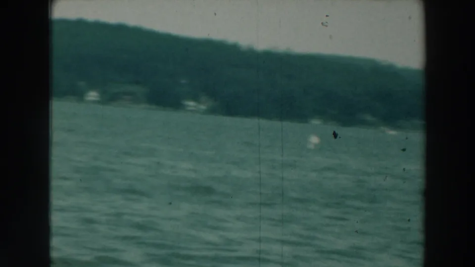 1964 8mm boat view on Lake Winnebago, with rippled water, sloping wooded shore, and small distant white shapes near the shoreline.
