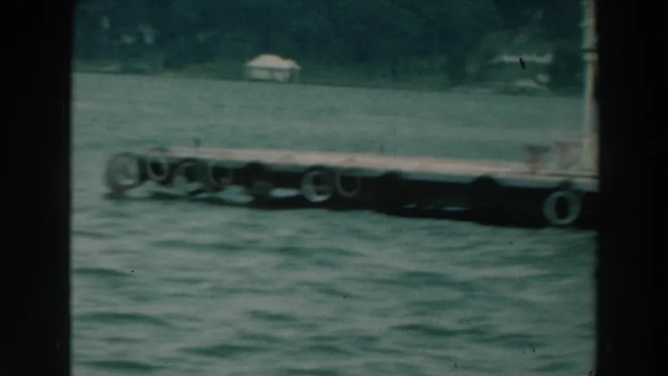 1960s 8mm frame of a floating dock with tire bumpers on Lake Winnebago, with wooded shore behind.
