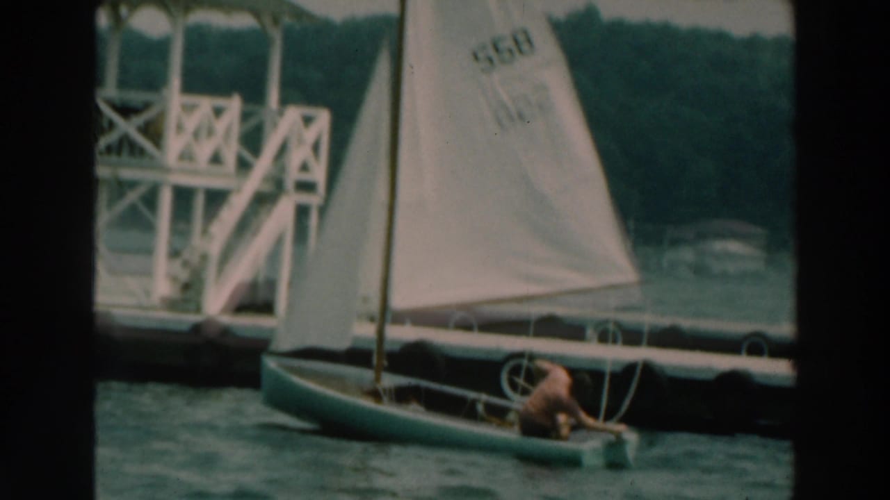 1964 Lake Winnebago 8mm frame showing a small sailboat with a shirtless sailor passing a white dock structure.