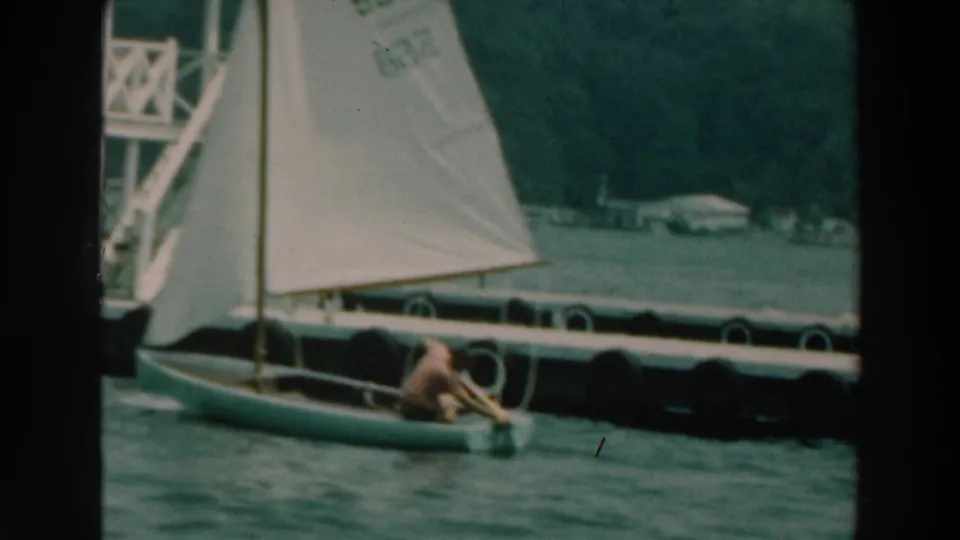 1964 Lake Winnebago frame with a small dinghy, numbered sail, floating docks, and tree-lined shore in the background.