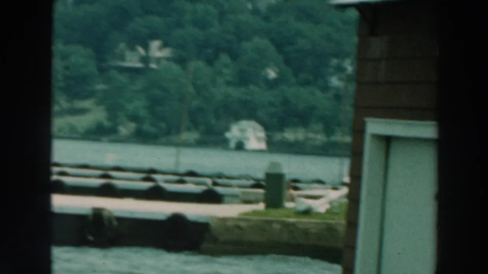 1964 Lake Winnebago 8mm frame with docks in the foreground, wooded shoreline across the water, and a building edge on the right.
