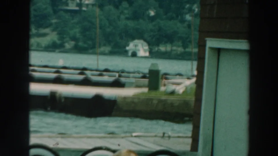 1964 Lake Winnebago home-movie frame looking past dock or boathouse framing toward water, trees, and a distant light-colored lakeside building.