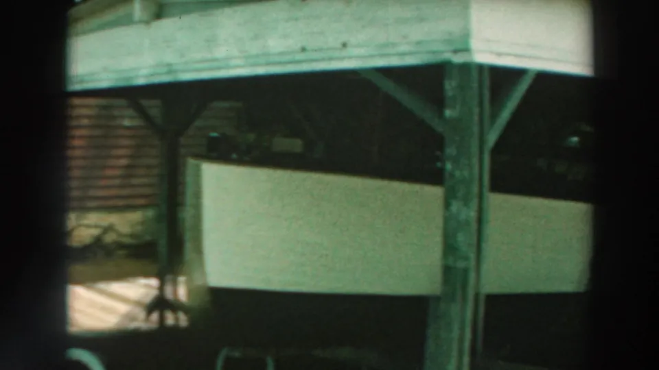 End frame showing the white boat stored below the pier, with beams, posts, and a small glimpse of shoreline or ground beyond.