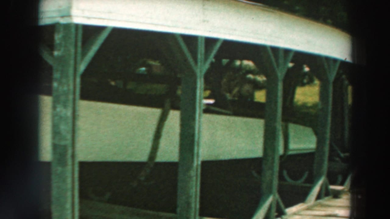 Boat hull beneath a roofed wooden shelter with posts and grass beyond.