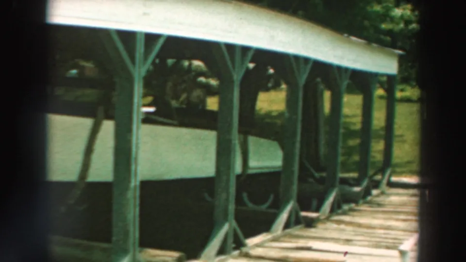 1964 Lake Winnebago home-movie view of a boat under a wooden lakeside shelter, with a plank walkway and sunlit grass nearby.