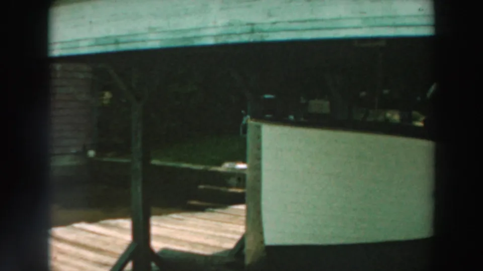 1964 Lake Winnebago frame showing the end of a sheltered boat, a wooden deck or walkway, and shaded grass beyond.