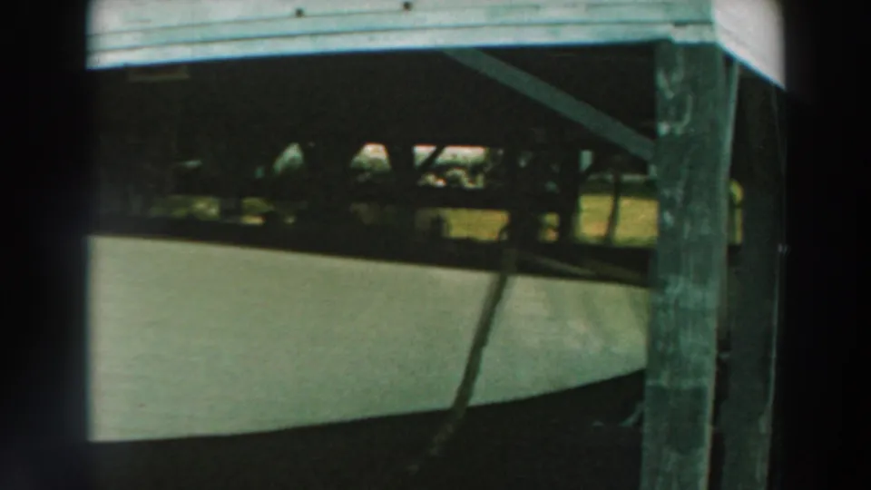 The camera frames the side of a white boat beneath the pier, with dark beams and posts around it.